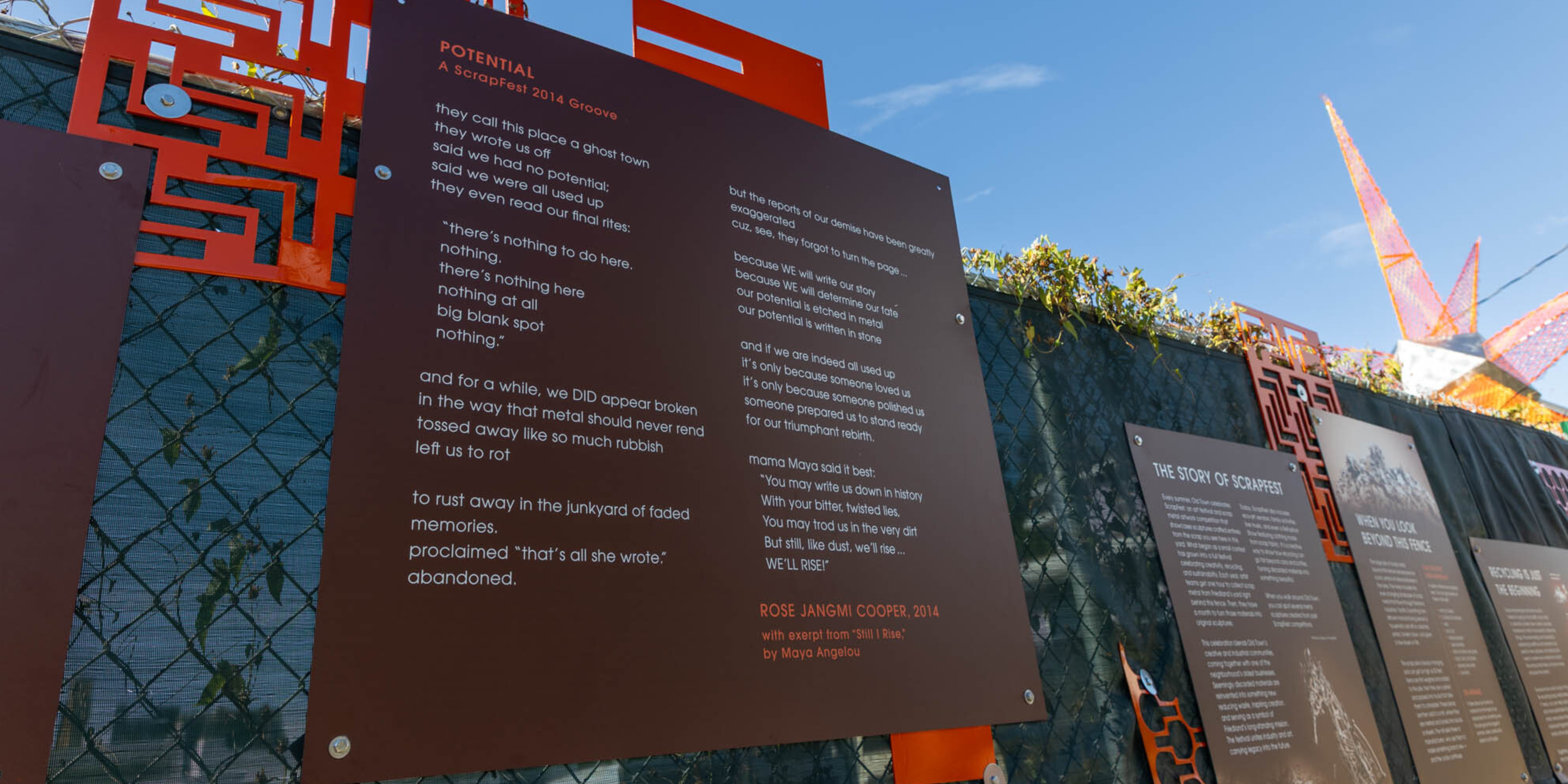 Boards for a scrap metal art installation display a poem titled "Potential" and pieces of scrap painted orange and displayed as art.